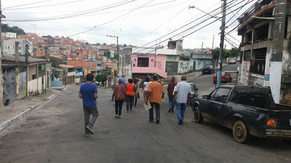 Pessoas caminham no meio de uma rua de bairro sem calçada, ao lado de carros