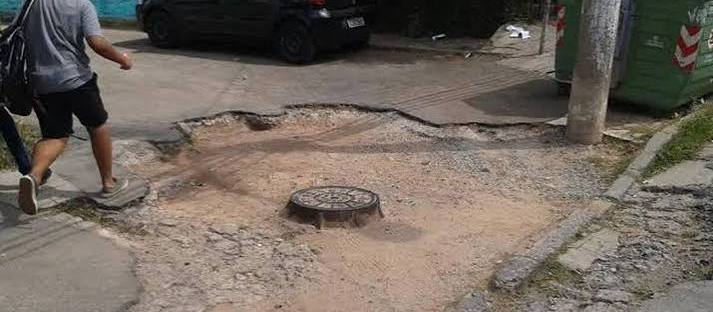 Pedestre caminha sobre calçada esburacada e quebrada, ao lado de carros estacionados